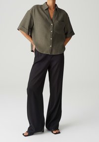 Chemise boutonnée kaki à manches courtes avec une poche poitrine, portée avec un pantalon noir à jambes larges et des sandales à enfiler noires.