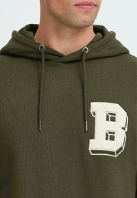 Hombre con sudadera verde olivo que tiene un parche de letra "B" blanca texturizada en el pecho, cuerdas visibles y una vista parcial de su cara.