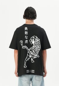 Schwarzes T-Shirt mit einem großen weißen Grafikdruck eines Tigers auf dem Rücken, begleitet von japanischem Text. Kurzärmlig, lässiger Schnitt, aus Baumwolle.