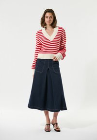 Maglione a righe rosse e bianche con scollatura a V e polsini a coste, abbinato a una gonna A-line in denim scuro con cuciture visibili. Sandali neri.