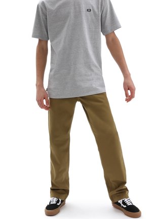 Mężczyzna w szarej koszulce Vans, brązowych spodniach i czarnych trampkach Vans stoi z nogami rozstawionymi na szerokość i rozluźnionymi ramionami.