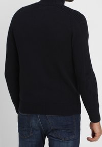 Námořnický svetr s vysokým límcem, žebrovanými detaily na ramenou a manžetách, přiléhavý design a hladká textura. Nosit s modrými džíny.