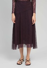 Jupe midi en dentelle de couleur violet profond avec des motifs floraux complexes, design en couches et un voile translucide ; accompagnée de sandales plates à lanières.