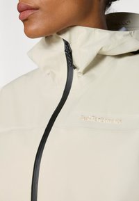 Gros plan d'une personne portant une veste beige Peak Performance avec un col montant et une fermeture éclair noire verticale.