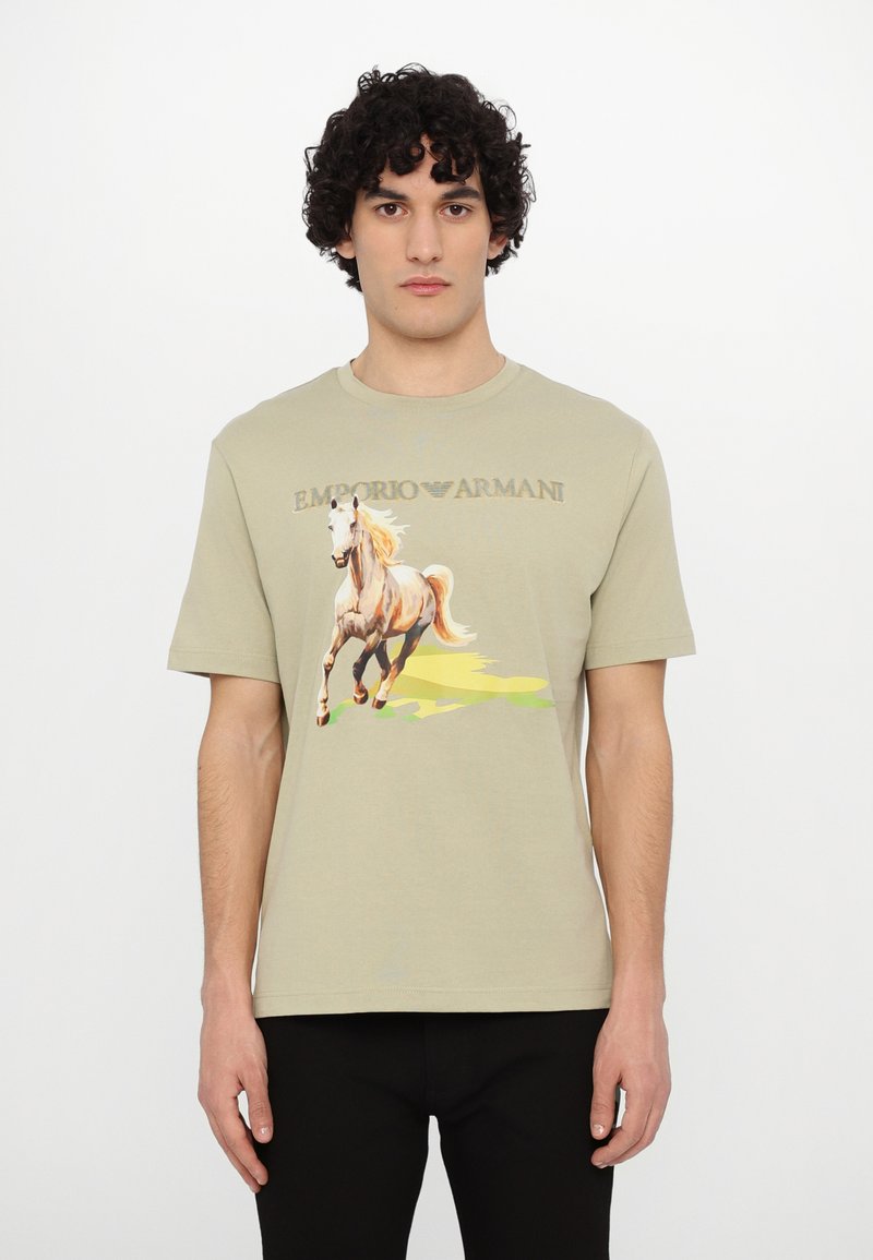 T-shirt en coton beige clair avec un graphique coloré de cheval et le texte "Emporio Armani". Manches courtes et col rond classique.