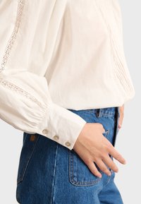 Weiße Baumwollbluse mit Spitzenverzierungen an der Schulter und einem gerüschten Ärmel mit drei Knöpfen am Bündchen, kombiniert mit Jeans.