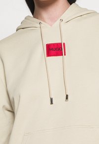 Felpa beige con cappuccio realizzata in cotone, dotata di un toppe rettangolare rossa con il testo "HUGO" e lacci con punte metalliche per la regolazione.