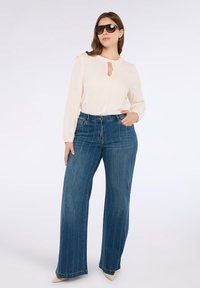 Blusa crema con dettaglio a chiave, abbinata a jeans di denim blu a gamba larga con cuciture verticali. Modella che indossa occhiali da sole oversize.