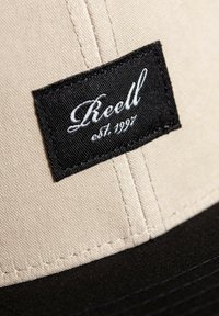 Gorra beige con visera negra, que presenta una etiqueta negra cosida que dice "Reell est. 1997" en texto blanco cursivo. Tela texturizada.
