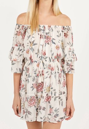Frau in einem schulterfreien weißen Kleid mit floralem Druck in Pink, Gelb und Blau, dreiviertellangen Rüschensämlen und tailliertem Schnitt mit Quasten.