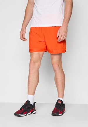 Lysorange sportsshorts med en glat tekstur, med en elastisk talje. Båret med sorte og røde sneakers og hvide sokker.