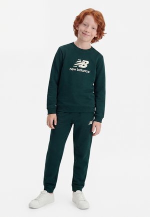 Junges rothaariges Kind steht und lächelt, trägt einen dunkelgrünen New Balance Pullover und passende Jogginghosen mit weißen Turnschuhen.