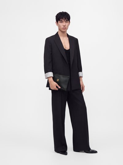 Νέος/Νεαρή που φοράει μαύρο oversized σακάκι και φαρδιά παντελόνια, κρατώντας μια μαύρη τσάντα clutch με χρυσή λεπτομέρεια σε απλό φόντο.