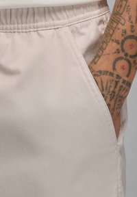 Lys beige stofshorts med elastisk talje, der har sidelommer og en glat tekstur. Synlig tatovering på armen i nærheden.