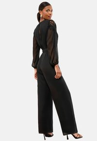 Mujer vistiendo un mono negro con mangas de encaje transparentes y pantalones de pierna ancha, de pie con la espalda vuelta, mirando sobre el hombro, usando sandalias de tacón.