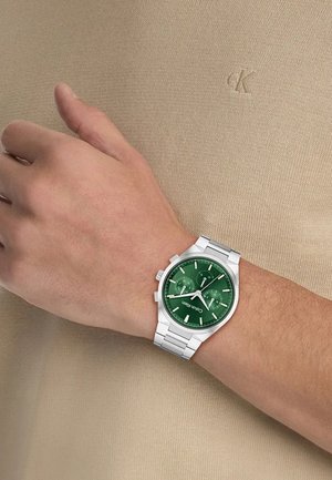 Montre à cadran vert avec bracelet en acier inoxydable argenté. Elle comporte trois sous-cadrans, des index heures argentés et un fond texturé kaki.