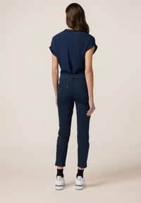 Navy blauwe top met korte mouwen en rugplooien, gecombineerd met tapered navy broeken. Model draagt witte schoenen met zilveren accenten en zwarte sokken.