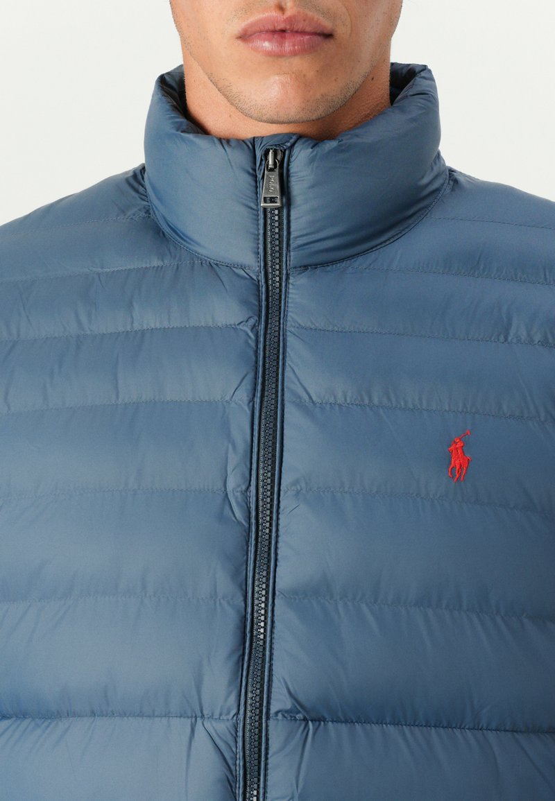 Chaqueta de plumas azul con un cuello alto, presenta un zipper frontal y costuras horizontales. Incluye un pequeño logo rojo en la parte izquierda del pecho.