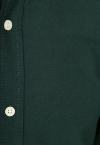 Dunkelgrünes Hemd aus strukturiertem Stoff, mit einer Reihe weißer Knöpfe an der Vorderseite und kontrastierenden Steppnähten.