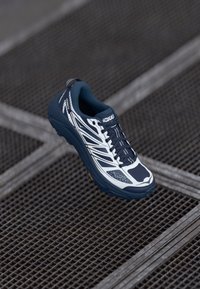 Modrá a bílá běžecká obuv s texturovaným svrškem, síťovinovými panely, polstrovanou podrážkou a hlubokými drážkami, s logem HOKA a reflexními detaily.