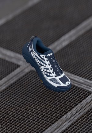 Modrá a bílá běžecká obuv s texturovaným svrškem, síťovinovými panely, polstrovanou podrážkou a hlubokými drážkami, s logem HOKA a reflexními detaily.