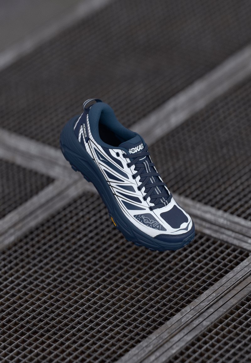 Modrá a bílá běžecká obuv s texturovaným svrškem, síťovinovými panely, polstrovanou podrážkou a hlubokými drážkami, s logem HOKA a reflexními detaily.