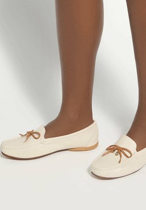 Dark-skinned legs wearing cream-colored leather moccasins with tan bow accents, standing on a plain white background.
