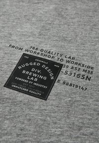 Šedá tkanina s texturovaným povrchem, obsahující černé obdélníkové logo a bílé textové informace o značce a designových specifikacích.