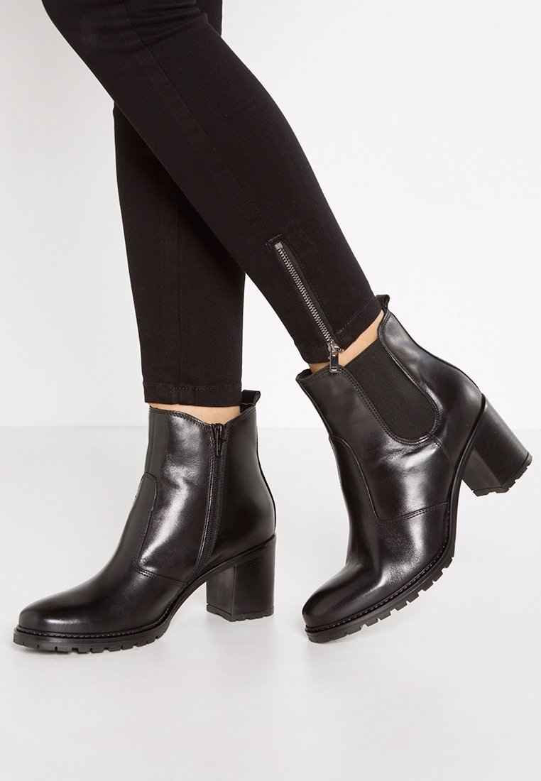 Jambes portant un pantalon slim noir avec des fermetures éclair latérales et des bottines en cuir noir à talons blocs sur un sol clair.