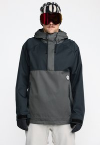 Veste pullover pour hommes en gris foncé et noir, avec une poche avant, une capuche ajustable et des fermetures à velcro aux poignets. Portée avec un bonnet à motifs et des lunettes de protection.