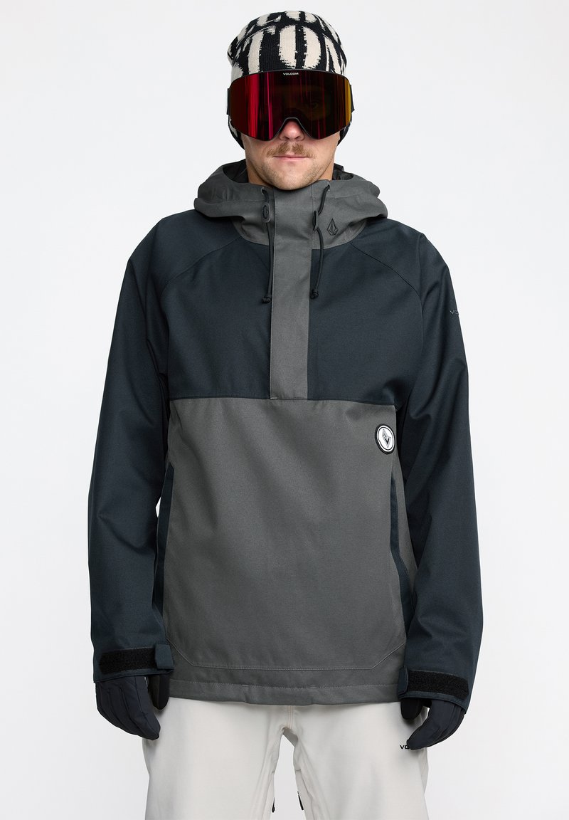 Veste pullover pour hommes en gris foncé et noir, avec une poche avant, une capuche ajustable et des fermetures à velcro aux poignets. Portée avec un bonnet à motifs et des lunettes de protection.