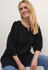 Čierna tunika s prepracovanými čipkovými rukávmi, výstrihom do V a voľným strihom. Mäkká textúra a detailný vzor na rukávoch.