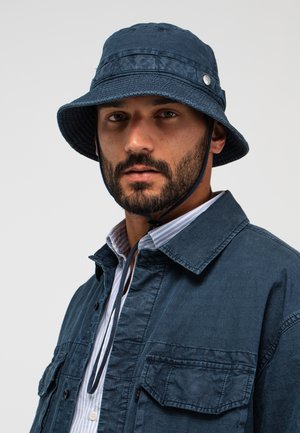 Mann mit dunklem Bart, der einen marineblauen Fischerhut mit Kinnriemen und eine passende marineblaue Jacke über einem weiß-blau gestreiften Hemd trägt.