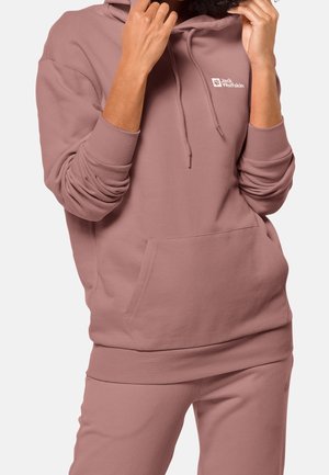 Personne portant un sweat à capuche mauve Jack Wolfskin et un pantalon assorti, ajustant les cordons de la capuche avec les mains près du visage.