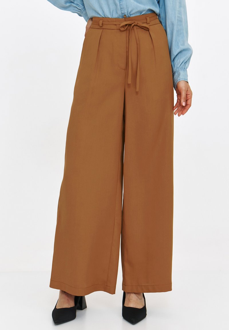 Brązowe spodnie z szerokimi nogawkami i paskiem w talii, z fałdami z przodu i gładką teksturą, w połączeniu z czarnymi butami na słupku.