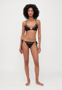 Woman with curly hair wearing black lace-trimmed bikini standing barefoot against a plain white background.