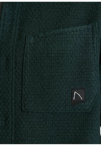 Zelená tkaninová bunda s textúrou, ktorá má náprsnú kapsu a logo štítok, s jemným vzorom tkania a mäkkým povrchovým dokončením.