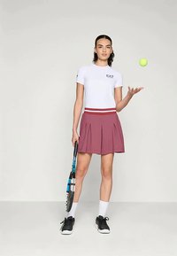 Balta prigludusi marškinėliai su juodu logotipu, derinami su plisuota tamsiai raudona sijonu. Juodi sportbačiai, balti kojinės ir raketė rankoje, mėgindamas sužaisti žaliu teniso kamuoliuku.