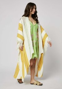 Keltainen ja valkoinen raidallinen kimono, jossa on fringed yksityiskohtia, käytetään vaaleanvihreän kukkakoristeisen mekon päällä. Avoin etuosa, löysä malli.