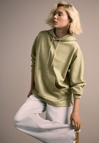 Voľná oversize mikina olivovo zelenej farby s predným vreckom a kapucňou na šnúrku, v kombinácii s voľnými svetlošedými nohavicami s textúrovanými detailmi.