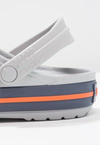 Grå Crocs med en marineblå base og en oransje stripe, med strukturert overflate og justerbare stropper. Signaturlogo på siden.