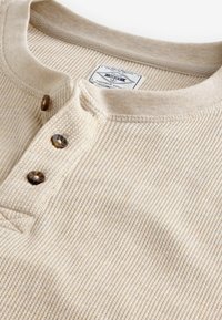 Hellbeige Henley-Shirt aus strukturiertem Material, mit rundem Ausschnitt, einer Knopfleiste mit drei braunen Knöpfen und einem gewebten Etikett im Inneren.