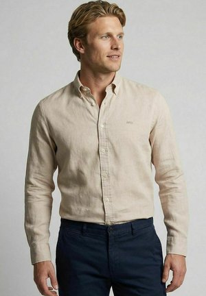Man with light brown hair wearing a beige button-up shirt tucked into dark navy pants, standing against a plain gray background.