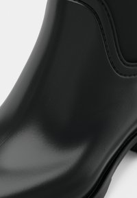 Botas pretas impermeáveis com um acabamento brilhante, apresentando uma ponta arredondada e uma textura suave. Os detalhes das costuras acrescentam interesse visual ao longo das bordas.