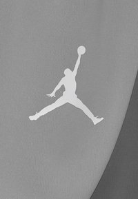 Sylwetka koszykarza skaczącego z szeroko rozstawionymi nogami i jedną ręką w górze trzymającego piłkę na fakturowanej tkaninie.