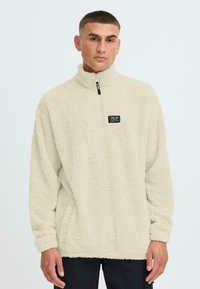 Hellbeiger Fleece-Pullover mit hohem Kragen, vorderem Viertel-Reißverschluss und strukturiertem Stoff. Verfügt über ein kleines schwarzes Markenlabel auf der Brust.