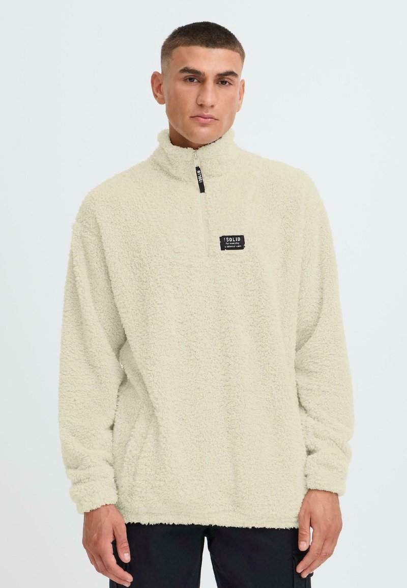 Hellbeiger Fleece-Pullover mit hohem Kragen, vorderem Viertel-Reißverschluss und strukturiertem Stoff. Verfügt über ein kleines schwarzes Markenlabel auf der Brust.