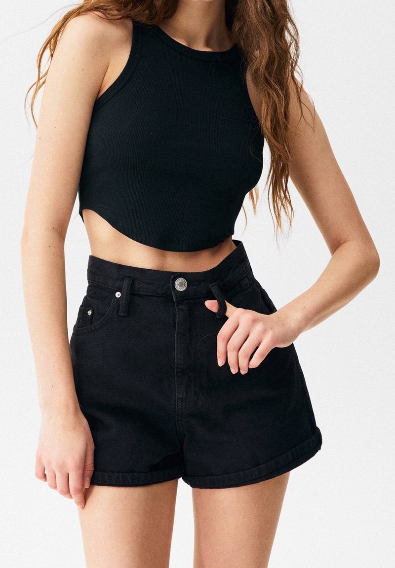 Černý žebrovaný crop top a černé džínové šortky s vysokým pasem a ohrnutými lemy, upevněné kovovým knoflíkem, zobrazené na horní části těla modelky.