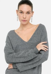 Pull en tricot gris avec décolleté en V, coupe ample et manches longues. Matériau texturé avec des éclats subtils. Modèle portant des boucles d'oreilles en chaîne argentée.