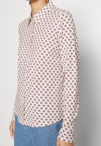 Camicia a maniche lunghe con bottoni di colore crema con motivo geometrico rosso. Presenta un colletto e un orlo dritto, realizzata in tessuto leggero.